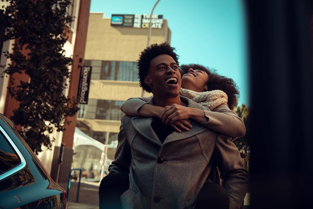 Happy couple outdoors: African American man carries woman piggyback in an urban setting, both smiling and looking up towards a bright sky.