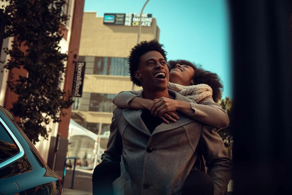 Happy couple outdoors: African American man carries woman piggyback in an urban setting, both smiling and looking up towards a bright sky.