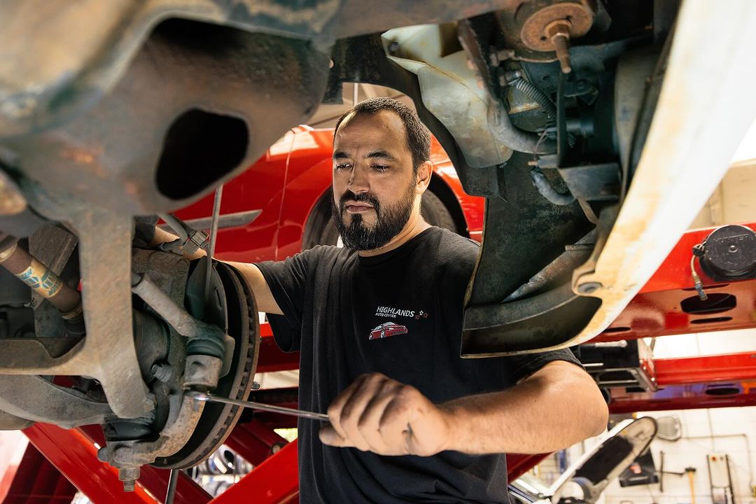 Man working on the undercarriage of a car, repairing the suspension, in a garage with automotive tools and equipment visible.