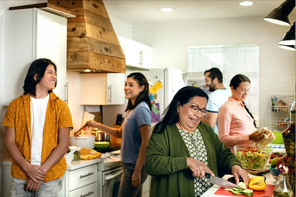 A diverse family group prepares food together in a brightly lit kitchen with white cabinets and a wooden range hood.