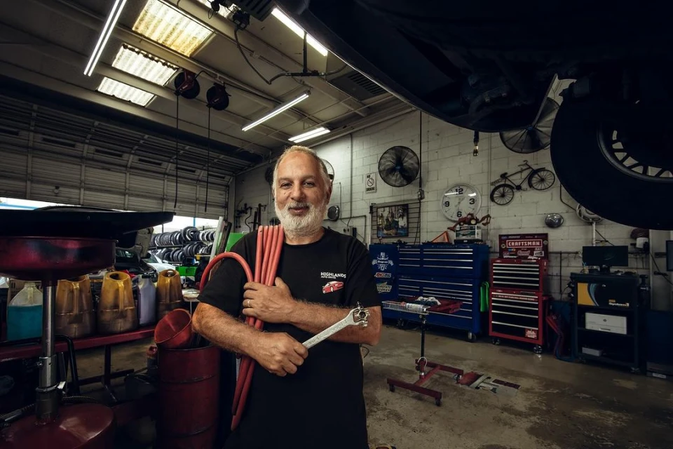 Adult male in a black t-shirt holding a wrench and red tubes, standing in an auto mechanic garage filled with equipment.
