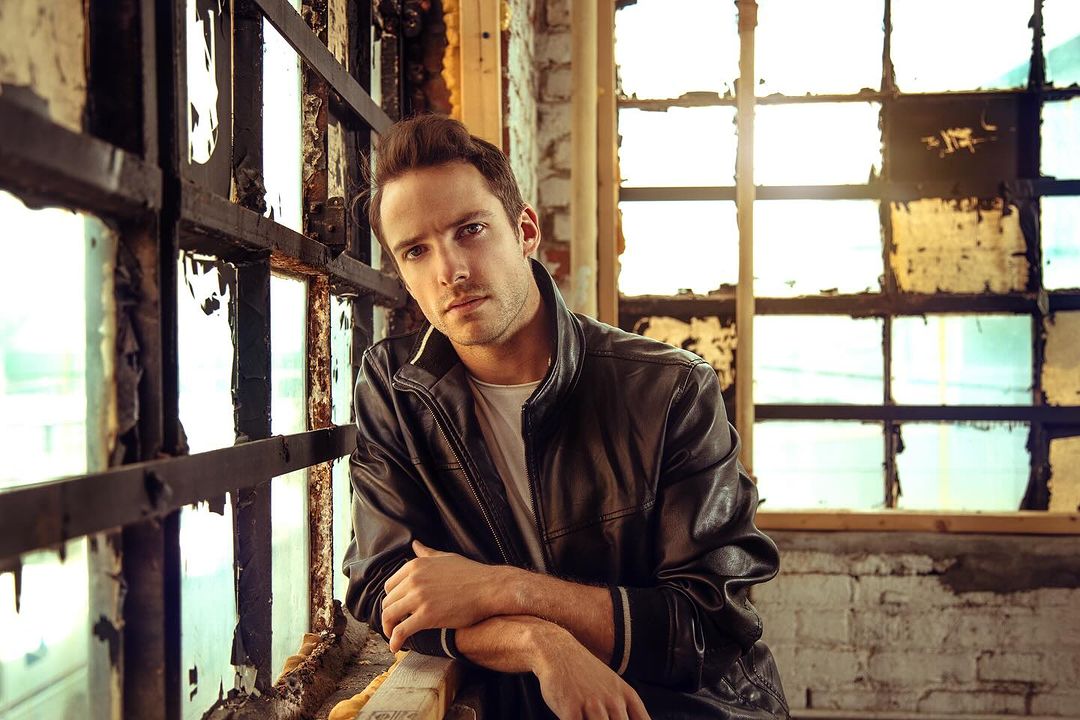 A portrait of a male model leaning against a window in a leather jacket, showcasing editorial fashion in an urban, indoor setting.