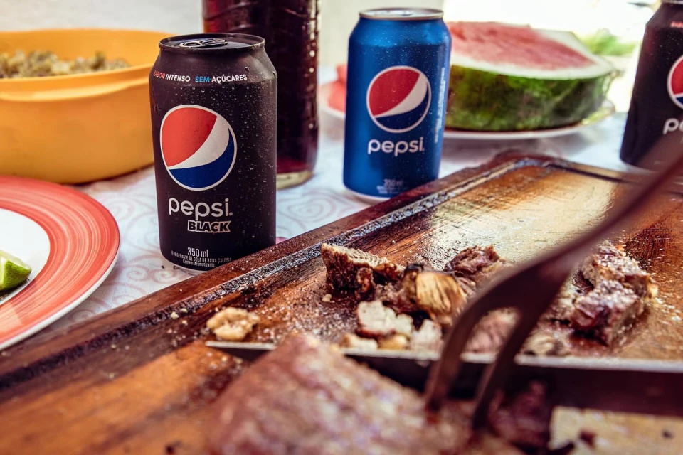 A picnic still life featuring Pepsi cans, sliced watermelon, a wooden board with food, and serving utensils on a white patterned tablecloth.