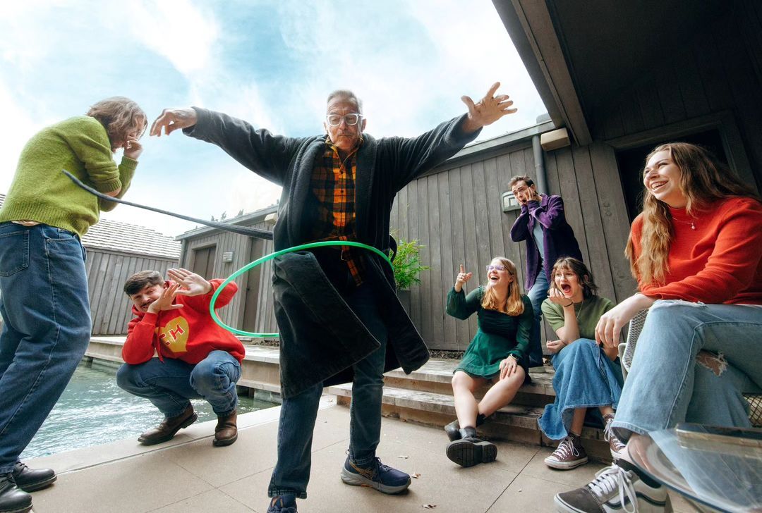 Group of eight people, including an adult male in a bathrobe using a hula hoop, gathered near a body of water with happy expressions.
