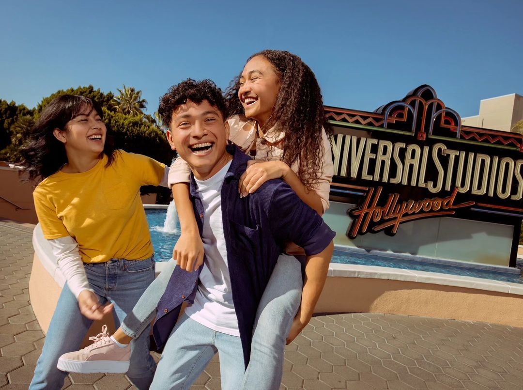 Three young adults laugh and pose in front of the Universal Studios Hollywood sign on a sunny day, creating a cheerful scene.