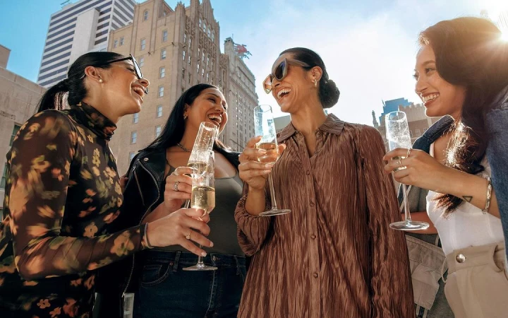 Four women laughing and toasting champagne in the city. They are dressed in stylish clothing as skyscrapers rise behind them.