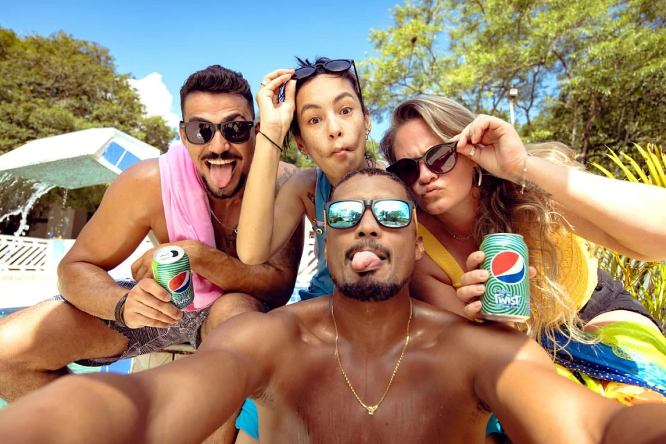 A group of four young adults posing for a selfie on a sunny day outdoors, holding cans of soda, wearing sunglasses and casual clothing.