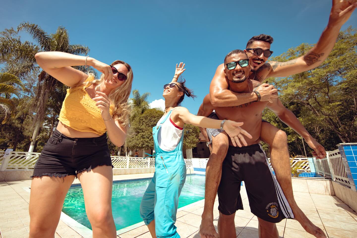 Four people pose near a pool on a sunny day, one giving a piggyback ride, creating a fun, summery vibe with trees and blue sky.