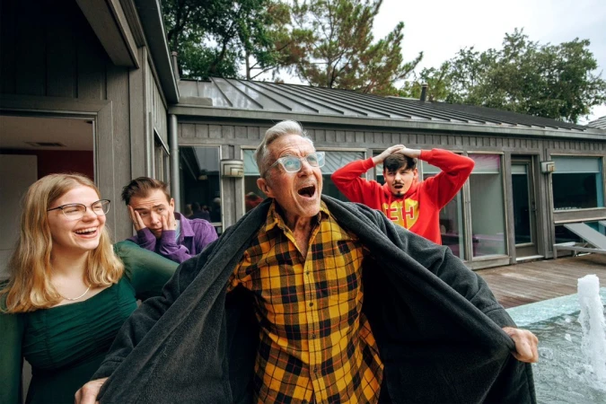 Outdoor shot of a group of four people with an elderly man in a yellow plaid shirt and grey robe standing in front of a water feature.
