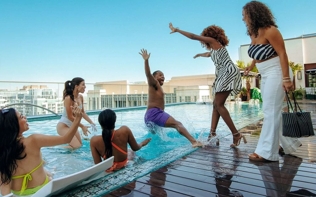 Group of six adults enjoying a rooftop pool party on a sunny day, with city buildings visible in the distance.