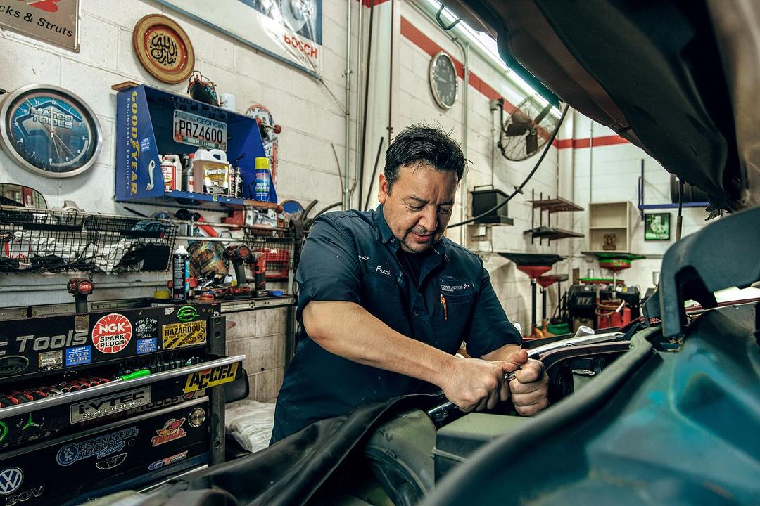 Auto mechanic working on a car in a repair shop, wearing a blue uniform, using hand tools, surrounded by tools and equipment, indoor setting.
