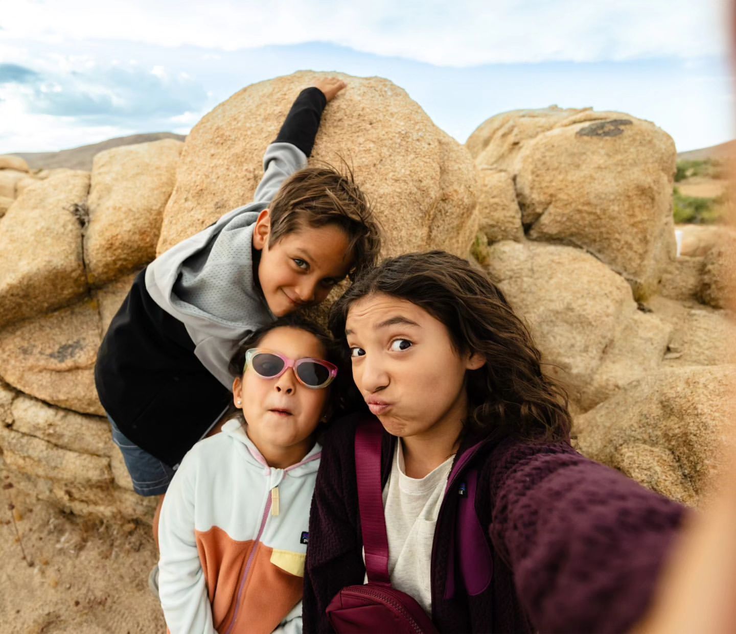 Three children pose for a selfie in a rocky, outdoor setting with a bright sky featuring light cloud cover, creating a cheerful, natural portrait.