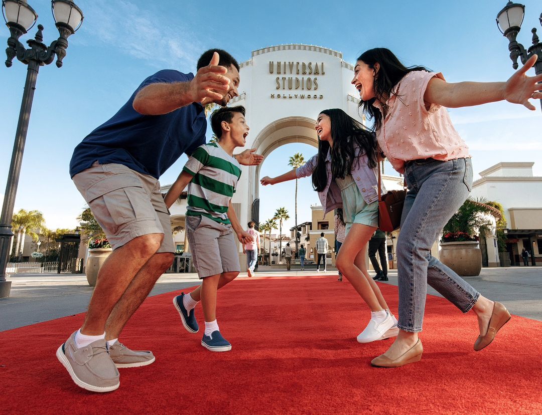 Family of four approaches Universal Studios Hollywood, arms outstretched in welcome, red carpet beneath feet, and clear blue sky above.
