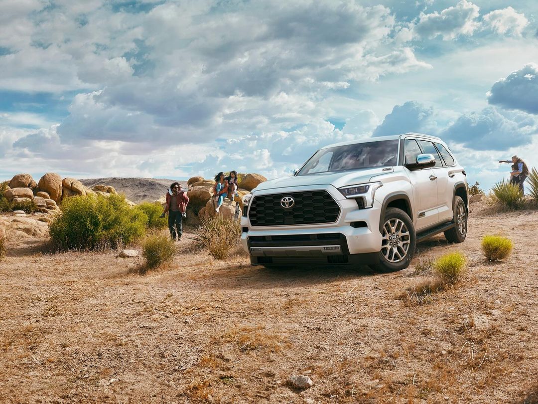 Silver Toyota Sequoia SUV parked on a desert plain with a family group nearby, under a cloudy sky. Automotive, family, outdoor theme.