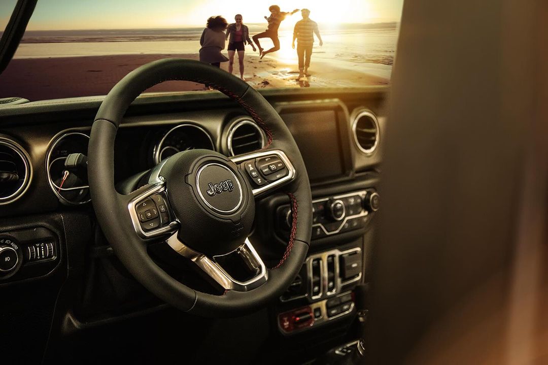 Car interior with a Jeep steering wheel in focus, family silhouettes enjoying a sunny beach day in the background, blurred.