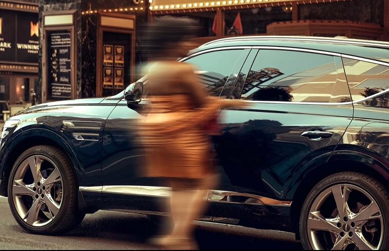 Dark blue SUV automobile parked on a city street near a theatre with a blurred person visible in the background, outdoors during the day.