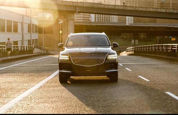 Sleek black Genesis SUV drives on a sunny urban road with a pedestrian jogging in the background, capturing motion and modern luxury.