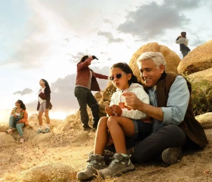Older man and young girl sitting on rock formations with family members exploring the landscape in the background
