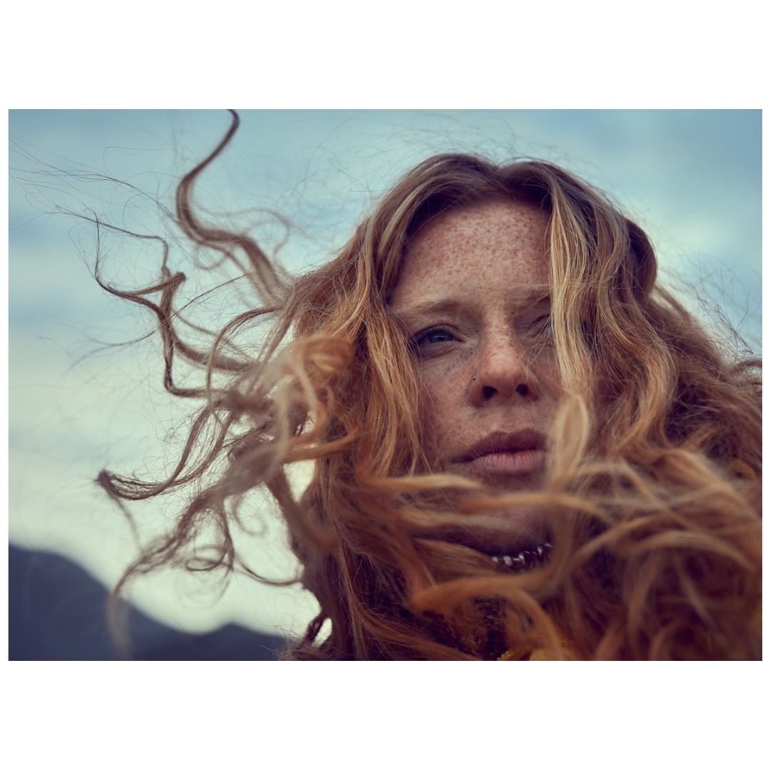 Close-up portrait of a red-haired adult woman with freckles and wind-blown hair, set against a backdrop of cloudy sky and distant mountains.