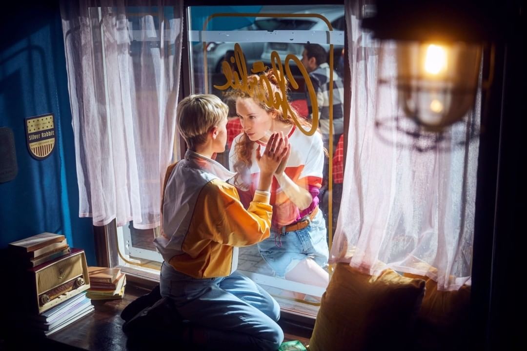 Two young women connect through a window, seen indoors with vintage decor and sunlight filtering through sheer curtains in the background.
