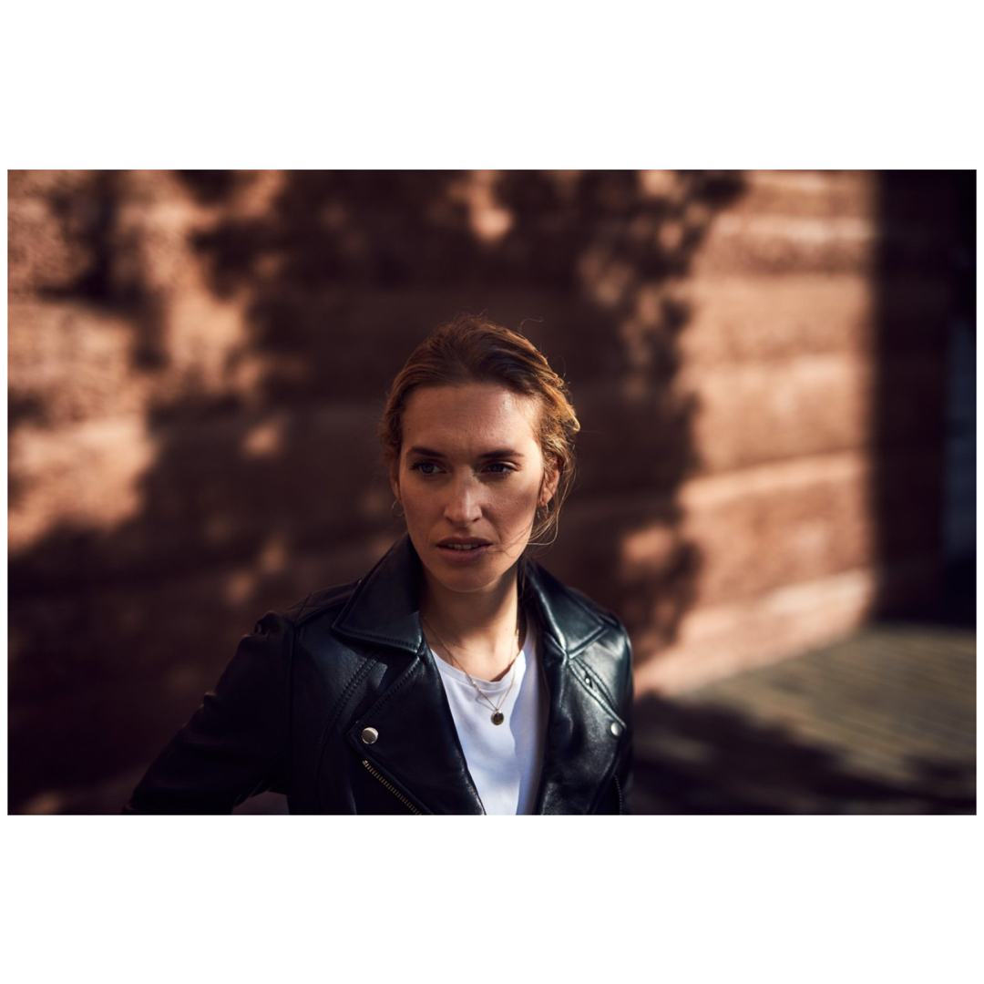 Woman in black leather jacket against blurred brick wall. Medium shot with shallow depth of field. Fashion, portrait, and lifestyle.