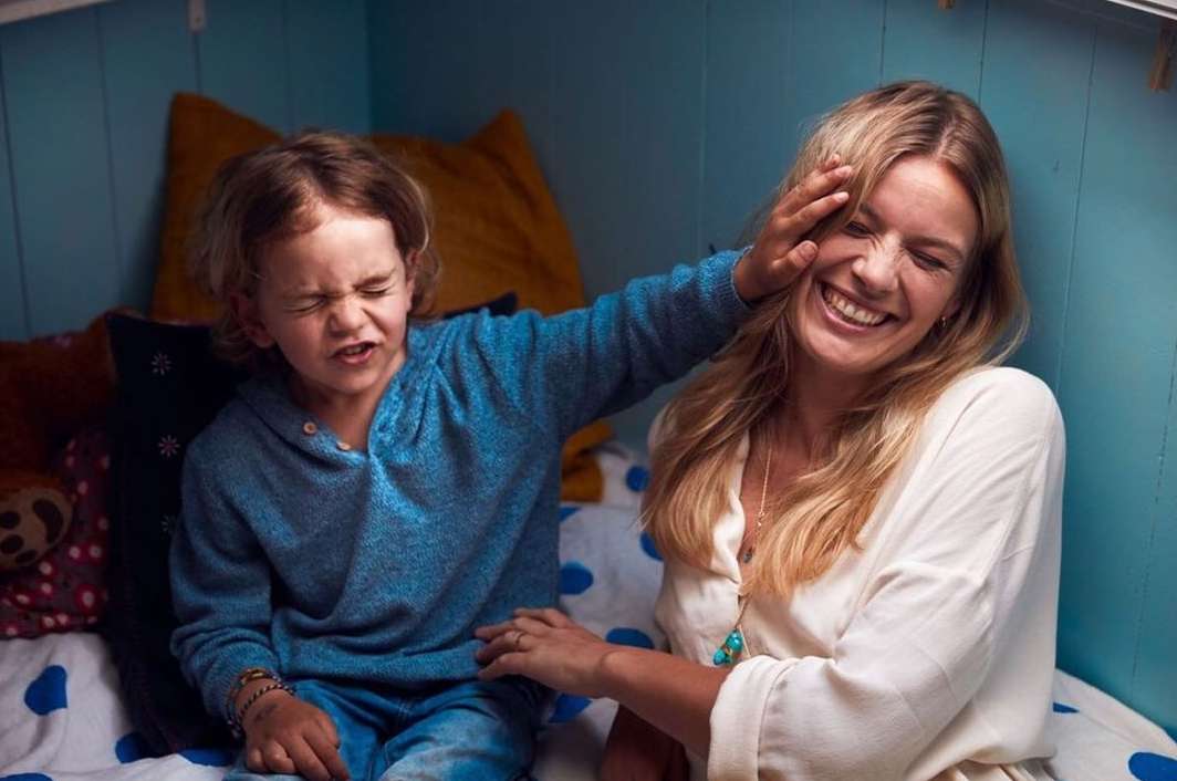 A blonde woman laughs as a boy with a blue sweatshirt covers her face with his hand indoors with a blue wall and patterned bedding.
