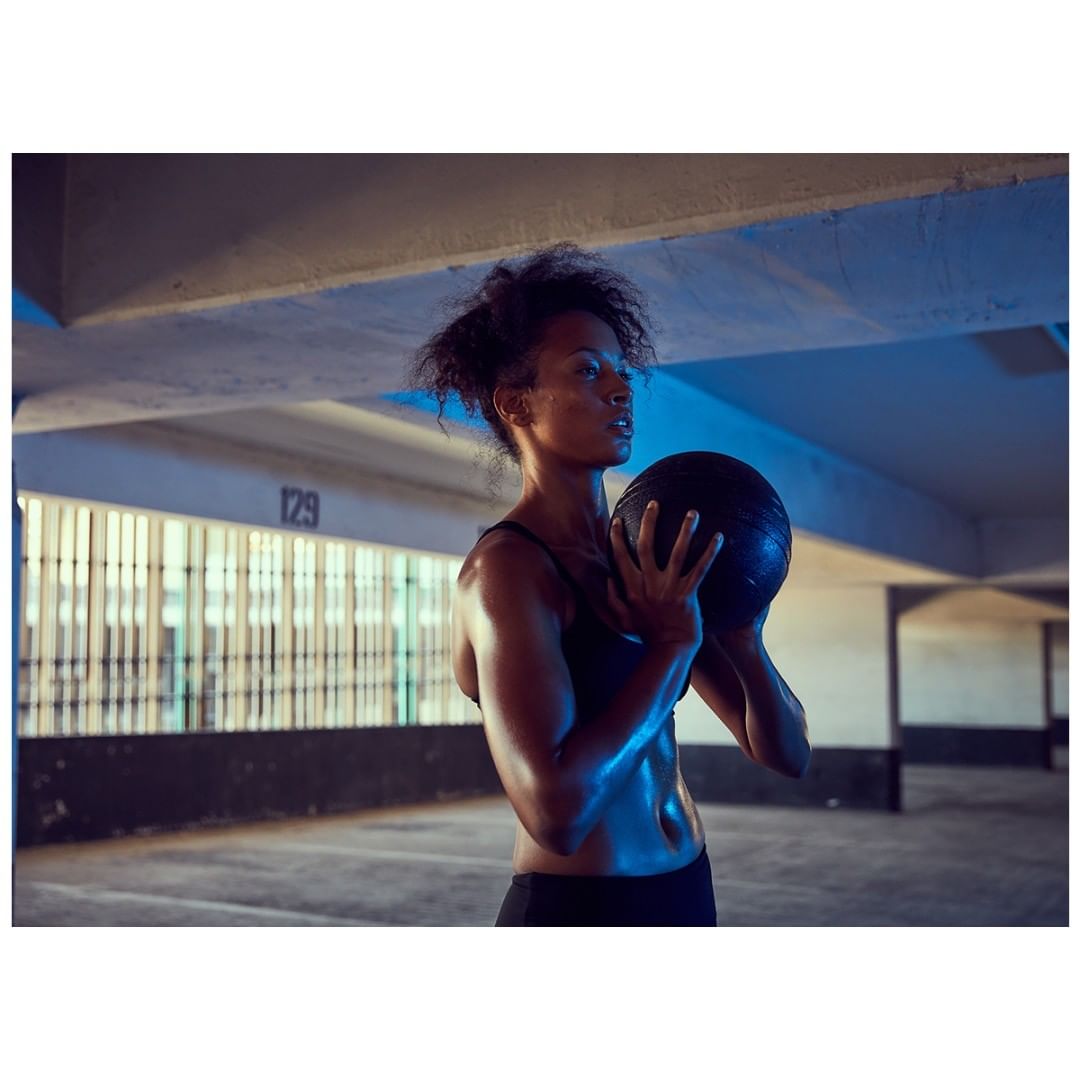 Athletic woman in sports bra holds a medicine ball in a dimly lit concrete parking garage. She is looking off to the side.