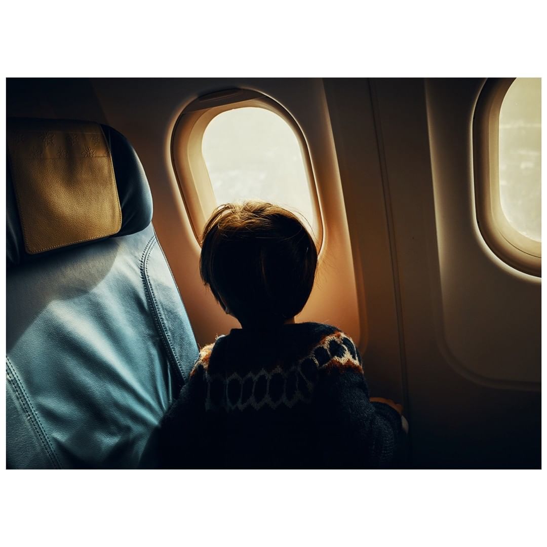 A child looks out of an airplane window during a flight. The interior of the plane is visible with the seat and window.
