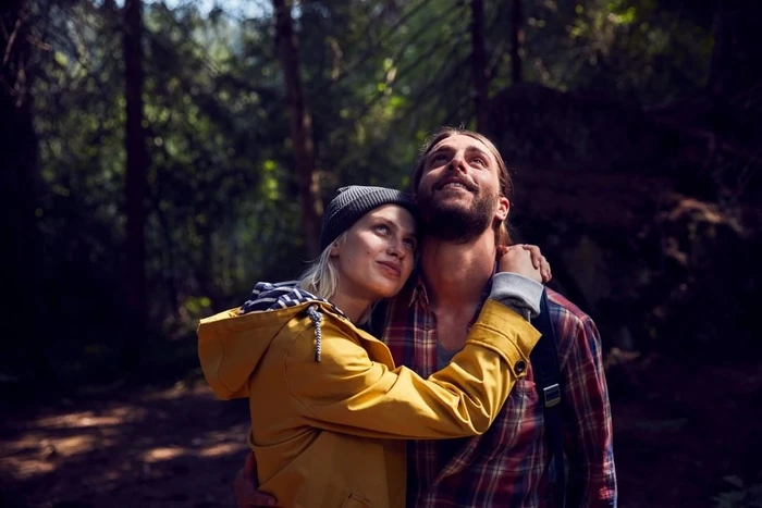 Man and woman hugging in a forest, looking up with joyful expressions while wearing outdoor clothing