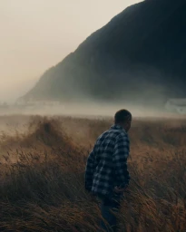 Man wearing a plaid shirt walking through a tall grass field towards misty mountains at dusk.