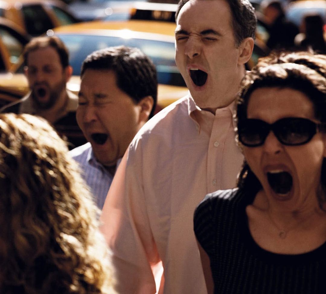 A group of adults, including men and women, yawns with their mouths open wide, possibly indicating tiredness or boredom in a candid moment.