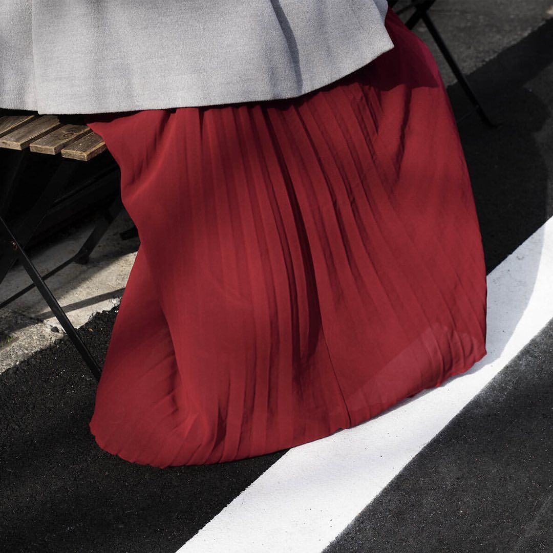 Close-up on a dark red pleated skirt draped over a wooden chair, with a grey jacket and a black and white asphalt background.