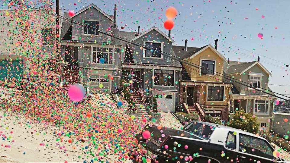 Colorful bouncy balls cascade down a San Francisco street, surrounding parked cars and homes on a sunny day in a whimsical scene.