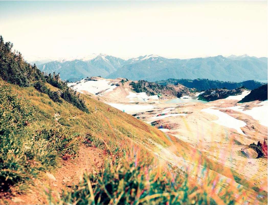 Scenic mountain landscape with a grassy slope in the foreground and snow-capped peaks in the distance under a light blue sky.