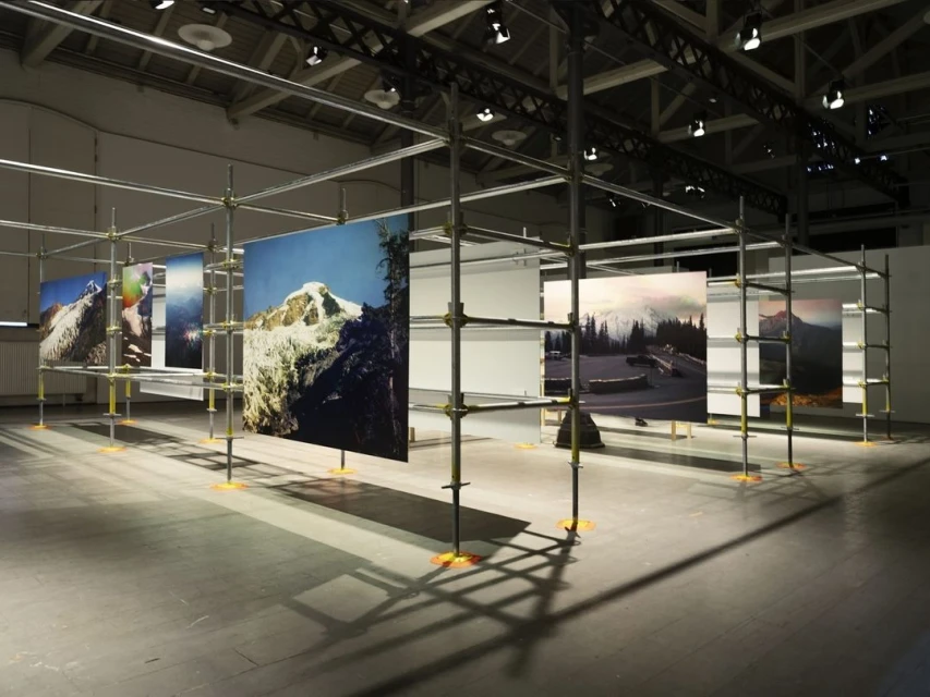 Artwork featuring photographs of snowy mountain landscapes hanging on a metal scaffolding structure in an indoor art gallery or exhibit space.