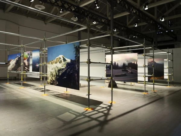 Artwork featuring photographs of snowy mountain landscapes hanging on a metal scaffolding structure in an indoor art gallery or exhibit space.