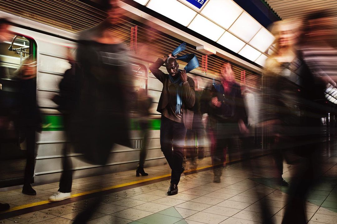 Blurred motion of diverse commuters at a subway station, a man in focus puts his scarf on with a train in the background.