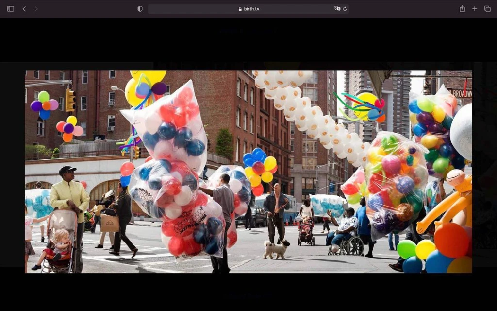 Street scene featuring people walking and in wheelchairs, surrounded by numerous colorful balloons in bags and garlands, city buildings backdrop.