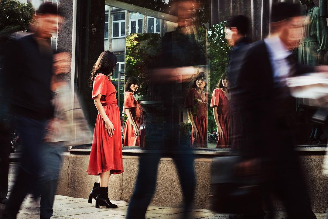 Woman in red dress stands outside shop window, reflected with blurred pedestrians passing by in fashion-forward urban scene.
