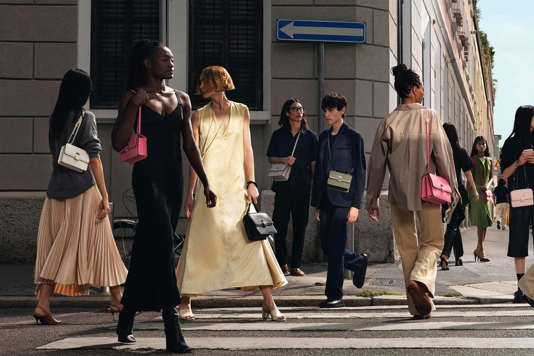 Several fashion models walk across a crosswalk with colorful purses in an urban setting during the daytime, promoting style and accessories.