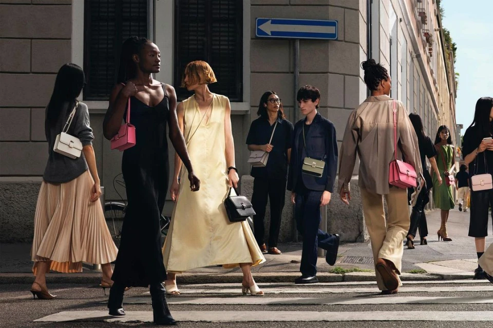 Several fashion models walk across a crosswalk with colorful purses in an urban setting during the daytime, promoting style and accessories.