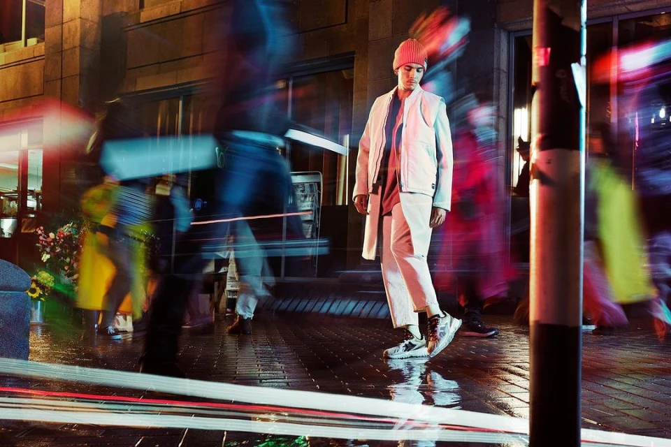 Male model in a beige jacket and matching pants walks on a wet city street at night with blurred pedestrians and car lights.