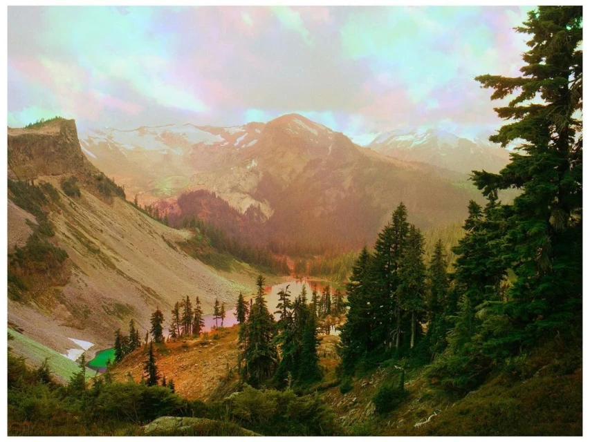 Scenic view of a mountain range with a lake, forest, and rugged cliffs on a hazy day with golden light and a partially cloudy sky.