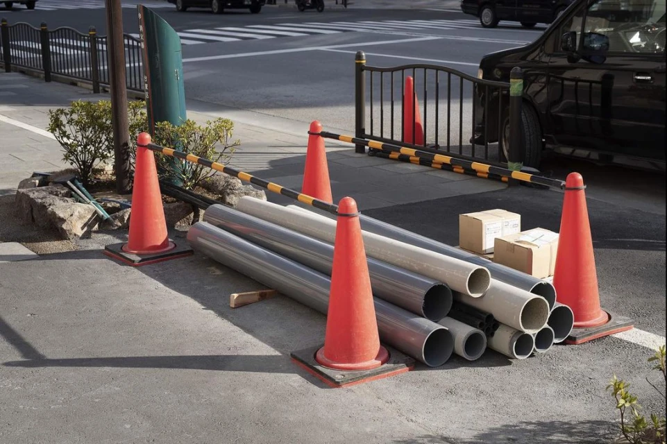 Traffic cones and gray industrial pipes near a black vehicle and sidewalk on a city street on a bright sunny day with natural light.