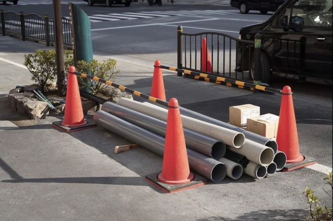 Traffic cones and gray industrial pipes near a black vehicle and sidewalk on a city street on a bright sunny day with natural light.