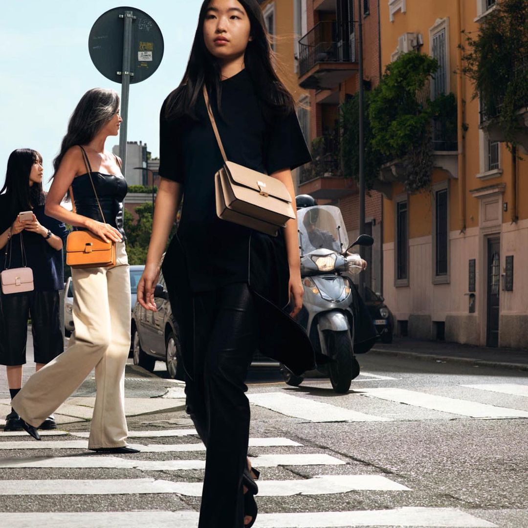 Three stylish women cross a city street on a sunny day. The woman in the foreground wears a black top and pants with a beige purse.