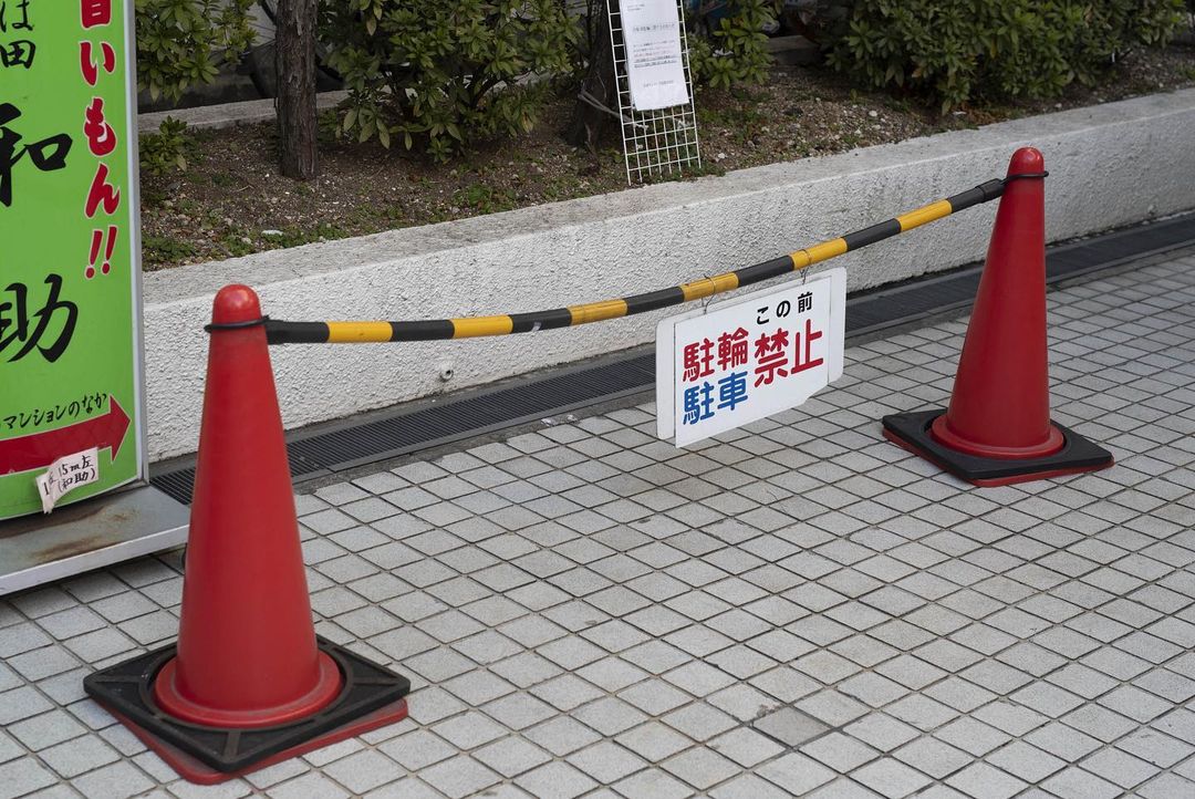 Two red traffic cones with a black and yellow striped pole barrier and a 'No Bicycle Parking' sign, set on a tiled outdoor surface.