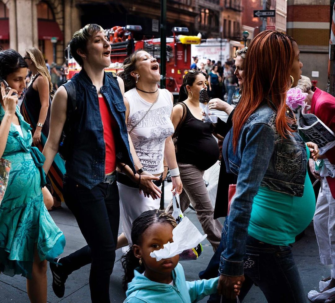 Candid street-style scene in New York City showcases a diverse group of pedestrians walking. Focus on a child with a tissue on her face.
