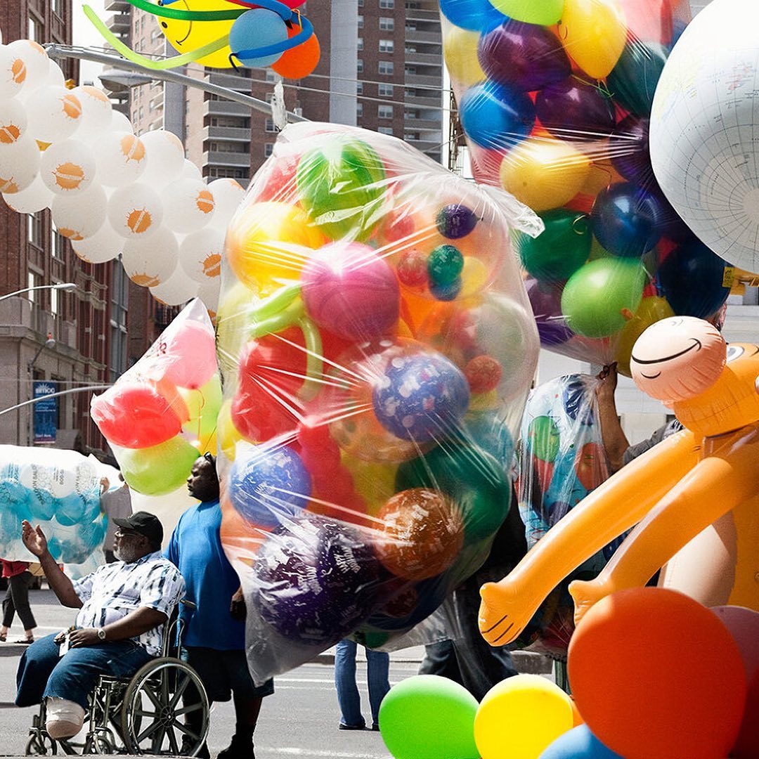 A man in a wheelchair sits next to a large bag of colorful balloons outdoors on a city street, surrounded by more balloons.