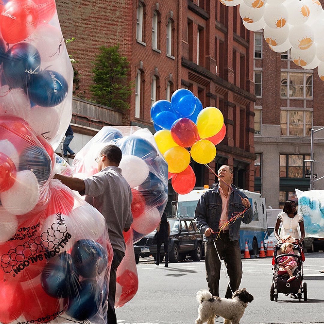 Street scene with vendors selling colorful balloons, a man walking a dog, and a woman pushing a stroller in an urban setting.